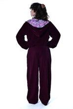Load image into Gallery viewer, A person shown from the back wearing a deep burgundy velvet hooded jumpsuit with a colorful paisley patterned collar, set against a plain white background. Buy now online from Emma's Emporium. Baggy unisex velvet jogger trousers with pockets. Emma's Emporium alternative hippie boho winter women's clothing. Alternative fashion, ethical, slow fashion for men and women.