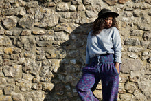 Load image into Gallery viewer, A woman wearing a light blue long-sleeve top, purple and blue patterned pants, and a black hat stands against a rustic stone wall. Buy now online from Emma's Emporium, soft warm fleecy winter hippie hippy trouser pants. Paisley pattern in blue or purple with pockets, unisex baggy trousers for men and women. Shop Emma's Emporium alternative fashion now, for ethical slow fashion, women's funky festival clothing.