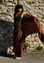 Load image into Gallery viewer, A woman stands outdoors against a rustic stone wall, wearing a dark olive green tank top, loose purple patterned pants, and holding a large matching purple shawl or wrap outstretched across the frame. Emma's Emporium paisley fleece blanket cardigan wrap throw. Shop online and buy now ethical slow fashion, unisex, men's, women's boho festival clothing.