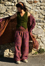 Load image into Gallery viewer, A woman stands outdoors against a rustic stone wall wearing a bohemian-style outfit with a dark green vest, wide-leg patterned pants in purple and orange, a matching headband, and a draped patterned shawl. Buy now online from Emma's Emporium, soft warm fleecy winter hippie hippy trouser pants. Paisley pattern in blue or purple with pockets, unisex baggy trousers for men and women. Shop Emma's Emporium alternative fashion now, for ethical slow fashion, women's funky festival clothing.