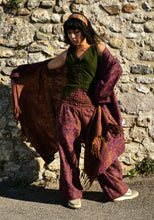 Load image into Gallery viewer, A person stands outdoors against a stone wall wearing loose bohemian-style clothing: an olive green sleeveless top, wide-leg mauve and purple patterned pants, and a matching draped shawl. The face is blurred. Emma's Emporium paisley fleece blanket cardigan wrap throw. Shop online and buy now ethical slow fashion, unisex, men's, women's boho festival clothing.