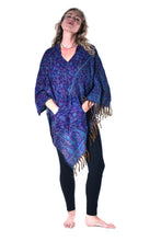Load image into Gallery viewer, A woman modelling a blue and purple paisley-patterned shawl with brown fringe, worn open over a pink top and black leggings, against a plain white background. Woman in an Emma's Emporium paisley poncho with a hood and front pocket. Shop Emma's Emporium online now, ethical slow alternative hippie festival fashion and women's clothing and apparel.