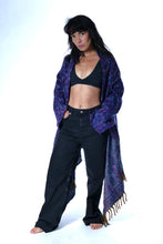 Load image into Gallery viewer, A woman modeling a long, open-front shawl with a purple and blue paisley pattern and fringe, worn over a black crop top and black wide-leg jeans against a white studio background. Emma's Emporium paisley fleece blanket cardigan wrap throw. Shop online and buy now ethical slow fashion, unisex, men's, women's boho festival clothing.