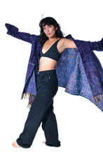 Load image into Gallery viewer, A woman posing with a large, open blue and purple patterned shawl or wrap draped over her arms, styled over a black bra top and dark wide-leg jeans against a solid white background. Emma's Emporium paisley fleece blanket cardigan wrap throw. Shop online and buy now ethical slow fashion, unisex, men's, women's boho festival clothing.