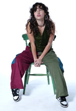 Load image into Gallery viewer, A woman with long curly dark hair sits on a green wooden chair against a white studio background. She is wearing a dark green sleeveless top, olive green wide-leg pants with blue patchwork details, black and white sneakers, and layered necklaces.
