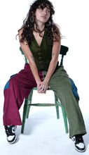 Load image into Gallery viewer, A woman with long curly dark hair sits on a green wooden chair against a white studio background, wearing a sleeveless olive green vest, wide-leg burgundy pants with blue patches, olive green wide-leg pants, and black-and-white sneakers.
