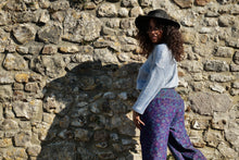 Load image into Gallery viewer, A woman wearing a black wide-brim hat, a light blue long-sleeve top, and purple patterned trousers stands in front of a rustic stone wall, looking back over her shoulder. Buy now online from Emma's Emporium, soft warm fleecy winter hippie hippy trouser pants. Paisley pattern in blue or purple with pockets, unisex baggy trousers for men and women. Shop Emma's Emporium alternative fashion now, for ethical slow fashion, women's funky festival clothing.
