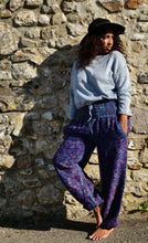 Load image into Gallery viewer, A woman stands barefoot against a rustic stone wall, wearing a light blue sweatshirt, dark purple and blue patterned jogger pants, and a black wide-brim hat. Buy now online from Emma's Emporium, soft warm fleecy winter hippie hippy trouser pants. Paisley pattern in blue or purple with pockets, unisex baggy trousers for men and women. Shop Emma's Emporium alternative fashion now, for ethical slow fashion, women's funky festival clothing.
