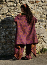 Load image into Gallery viewer, A woman stands with her back to the camera in front of a rustic stone wall, wearing a loose, patterned purple and orange shawl draped over her shoulders and matching patterned pants. Emma's Emporium paisley fleece blanket cardigan wrap throw. Shop online and buy now ethical slow fashion, unisex, men's, women's boho festival clothing.
