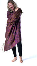 Load image into Gallery viewer, A woman modeling a large patterned shawl wrap in purple, burgundy, and gold tones with fringed edges, paired with black leggings, against a plain white background. Emma's Emporium paisley fleece blanket cardigan wrap throw. Shop online and buy now ethical slow fashion, unisex, men's, women's boho festival clothing.
